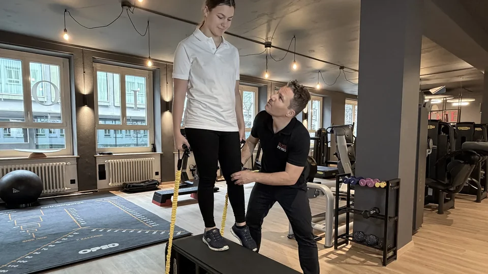 Physiotherapeut unterstützt eine Patientin am Knie bei einer Beinübung mit Widerstandsbändern auf einer Trainingsbank