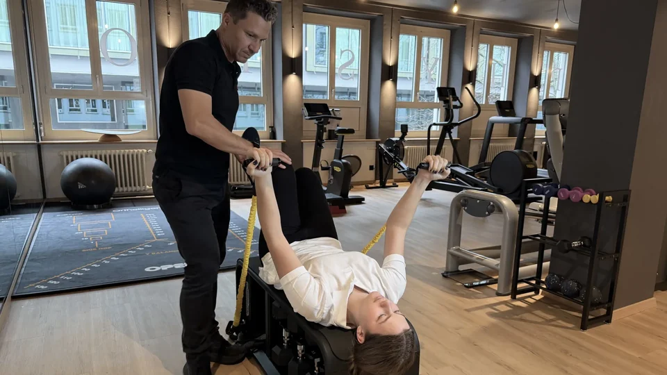 Physiotherapeut begleitet eine Patientin bei einer Schulterübung mit Widerstandsbändern auf einer Trainingsbank